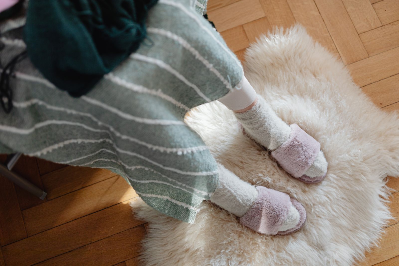 Overhead view of pink faux fur slide slippers worn with fluffy socks on a white sheepskin rug over parquet flooring