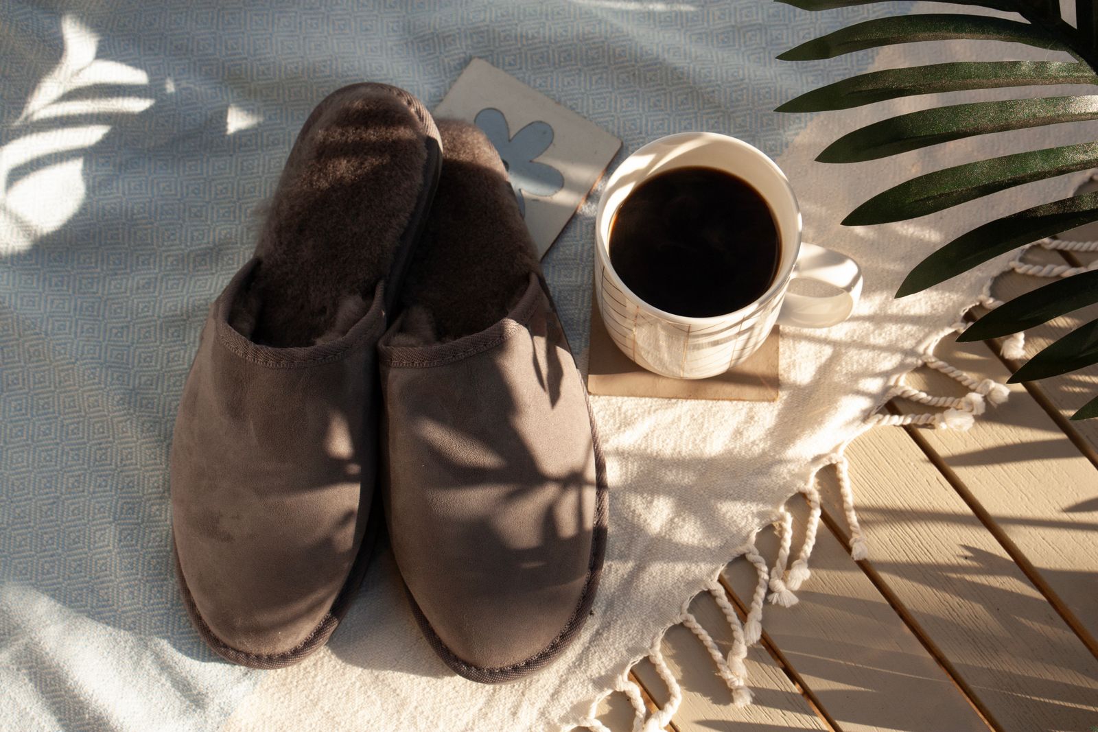 Gray suede scuff slippers with dark brown shearling lining beside a mug of black coffee on a sunlit outdoor blanket