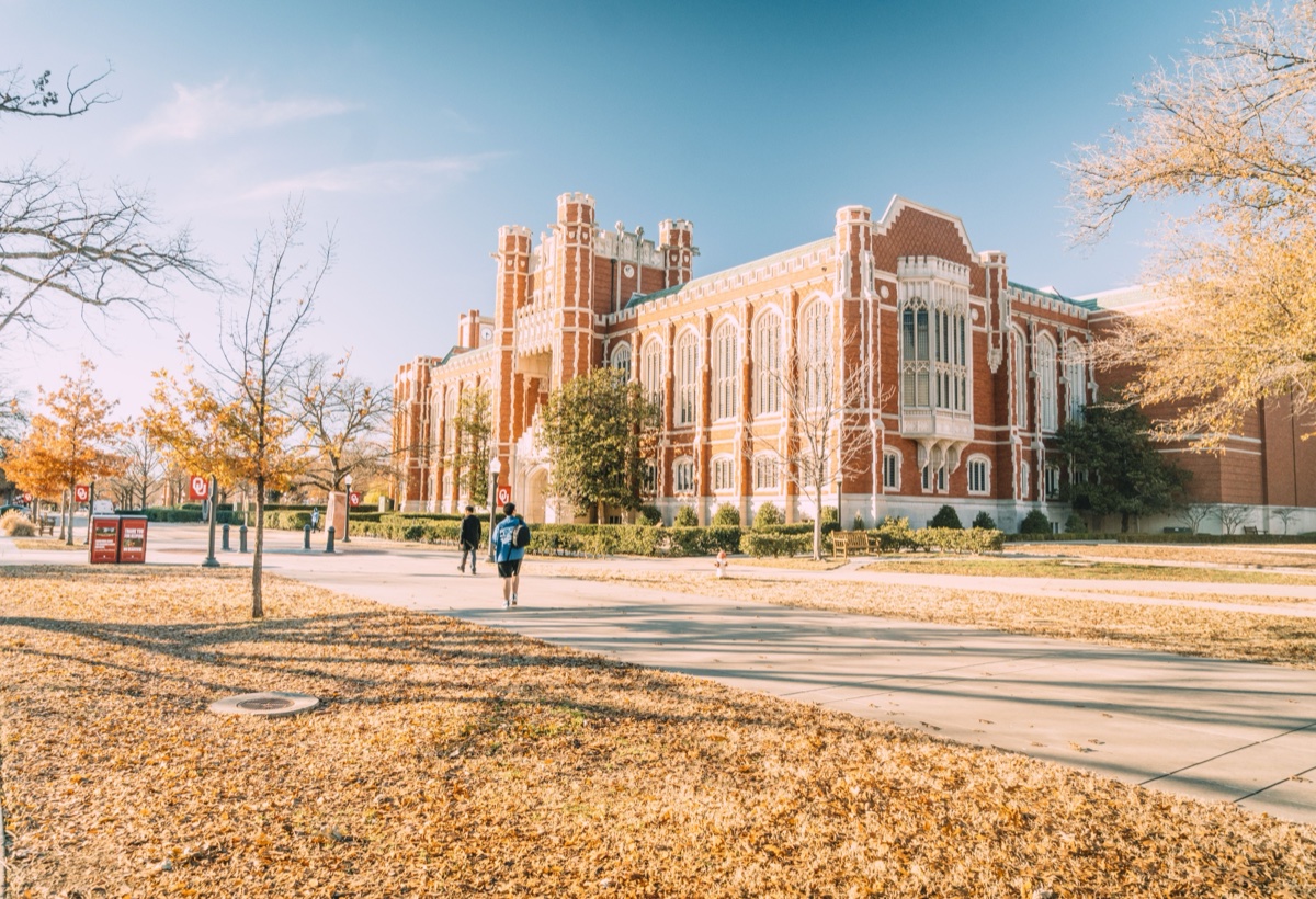 Students walking past a red-brick collegiate building on a leaf-covered campus path in autumn light