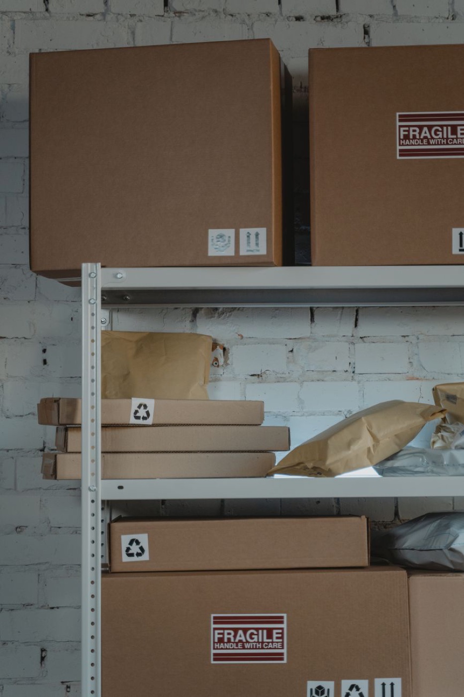 Cardboard boxes and padded mailers stacked on a metal warehouse shelf against a white painted brick wall