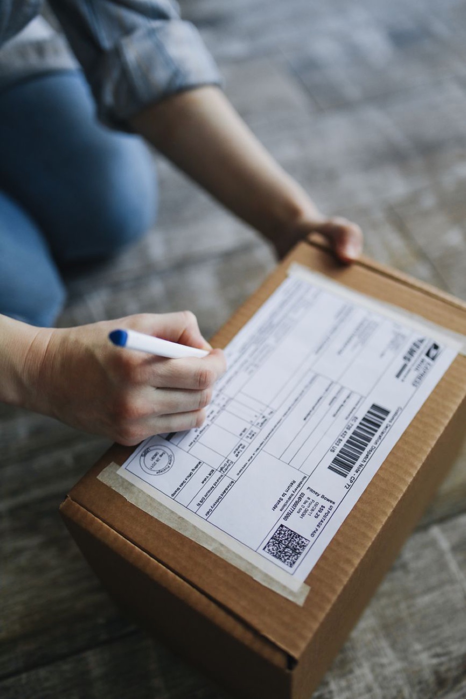 A person kneeling on a wooden floor writing on a printed postage label taped to a small cardboard box