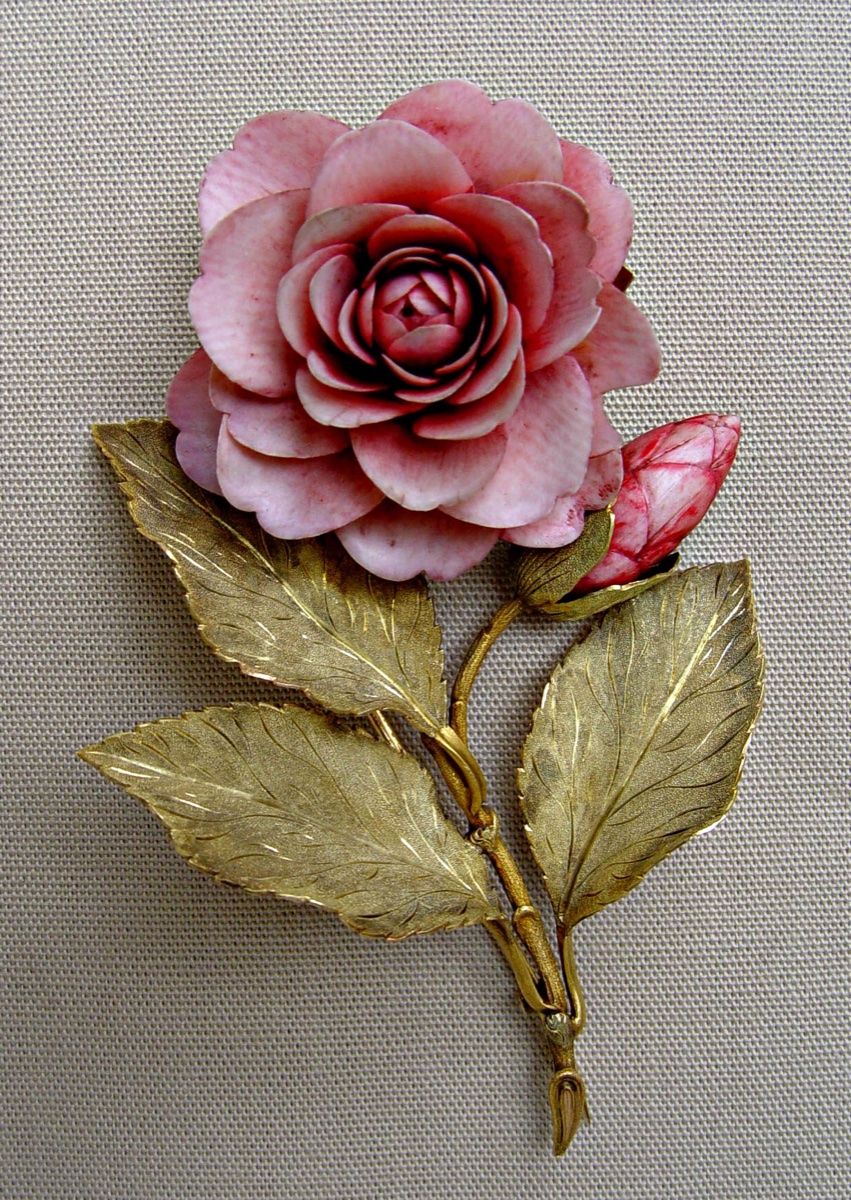A Victorian brooch in the form of a pink camelia with finely carved tinted petals and engraved gold leaves, photographed against beige fabric