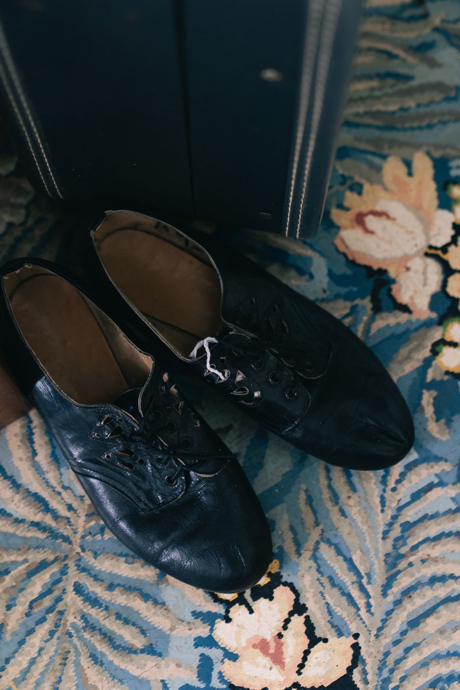A pair of worn black leather lace-up flats on a floral carpet next to a vintage suitcase