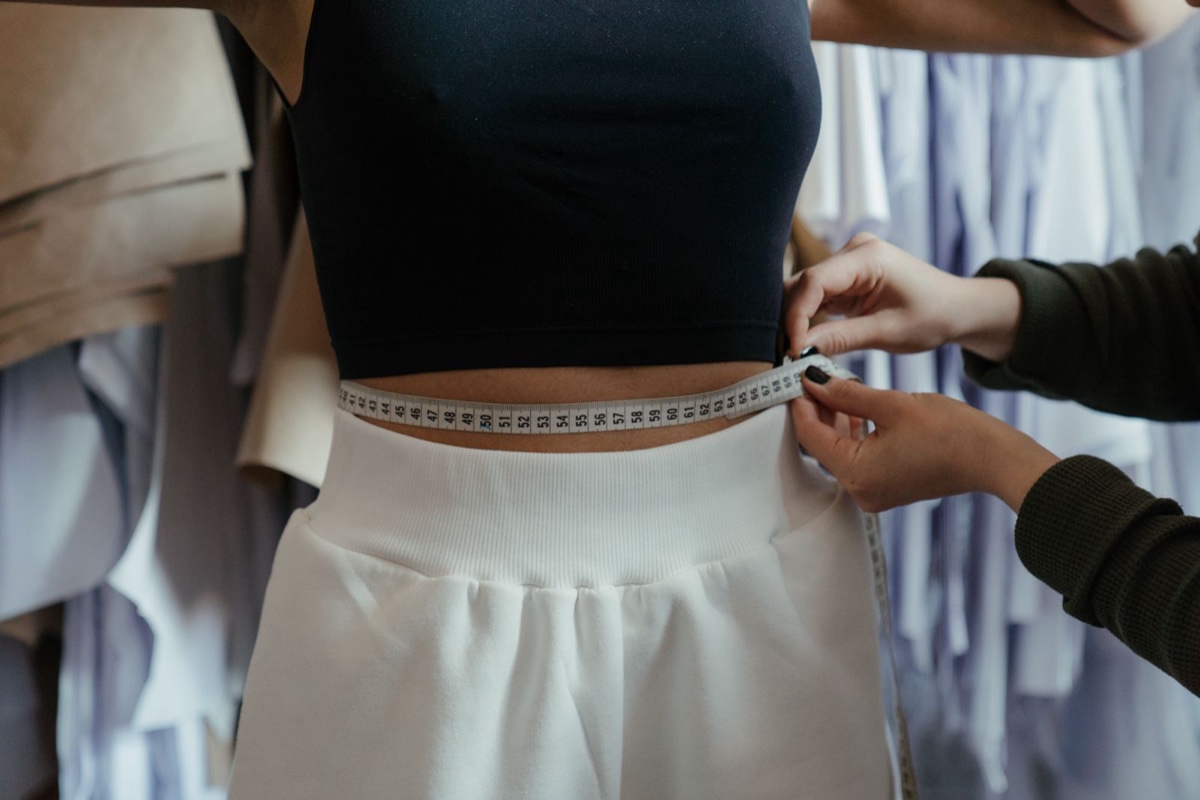 Hands holding a centimetre tape measure around a woman's waist in a fitting room, numbers in the 50s and 60s visible
