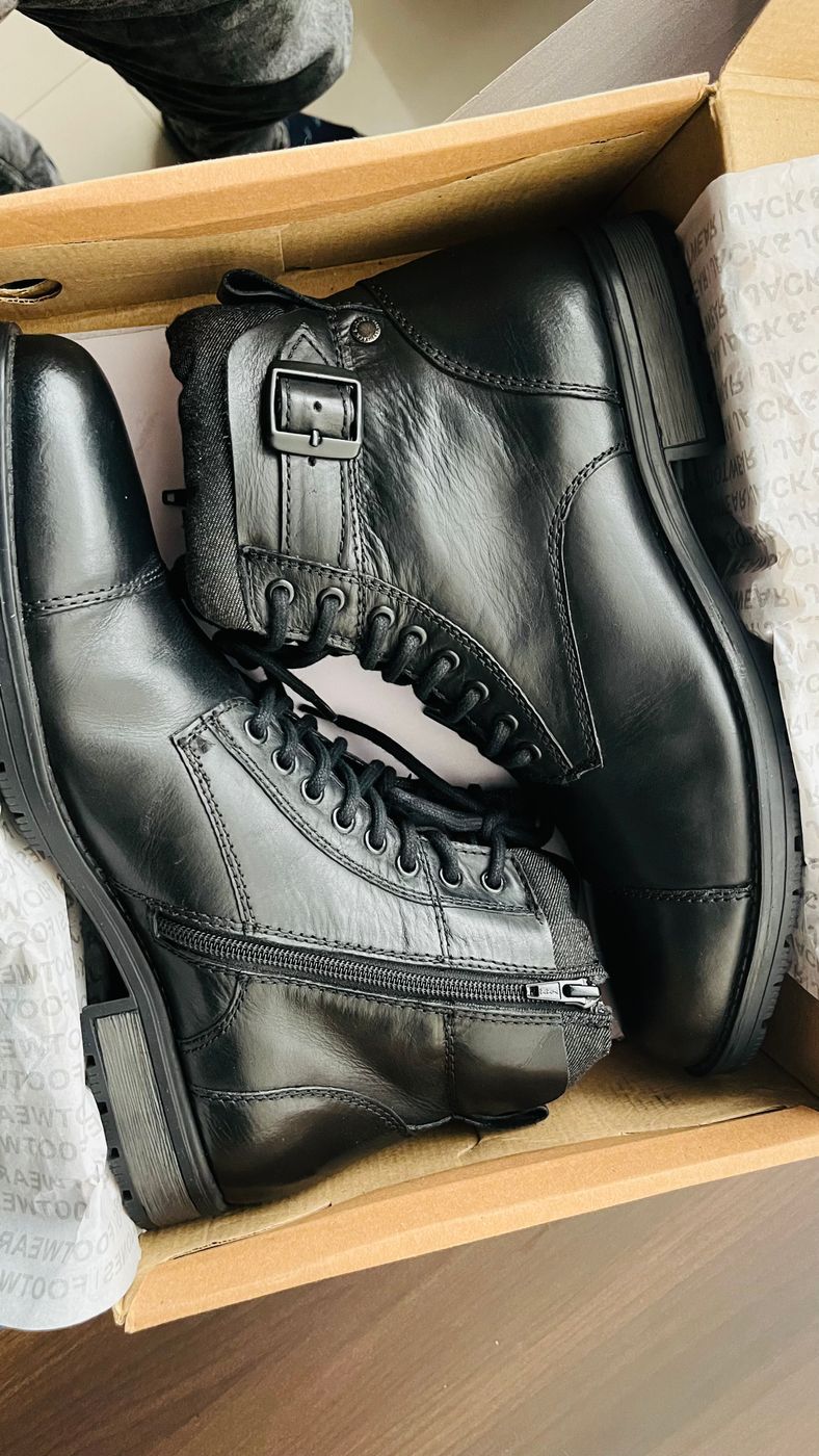 A pair of black leather lace-up boots lying in an open cardboard shoe box on a wooden floor