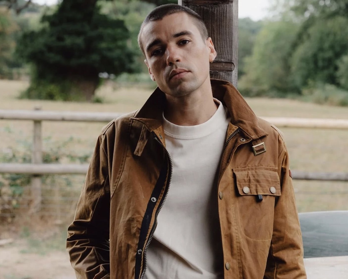 A tan waxed-cotton jacket, sheen showing at the creases, over a plain white tee by a paddock fence