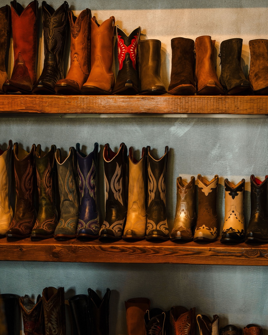 Rows of western boots in browns, black and cream lined up on three rustic wooden shelves against a pale wall