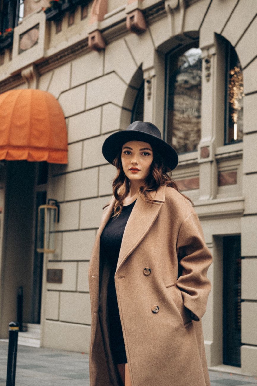 A woman in a loose double-breasted camel coat over a black top and black fedora, standing on a narrow European street with a cream stone facade behind her