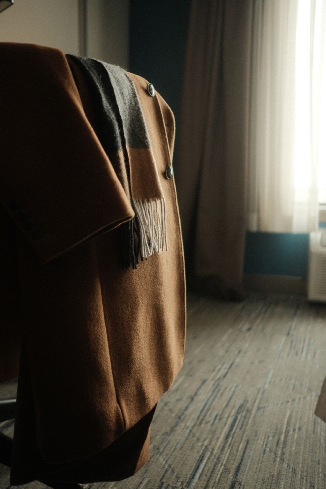 A deep tobacco-brown camel coat draped over a chair in a dim hotel room, a striped wool scarf hanging over its shoulder, daylight coming through sheer curtains behind it
