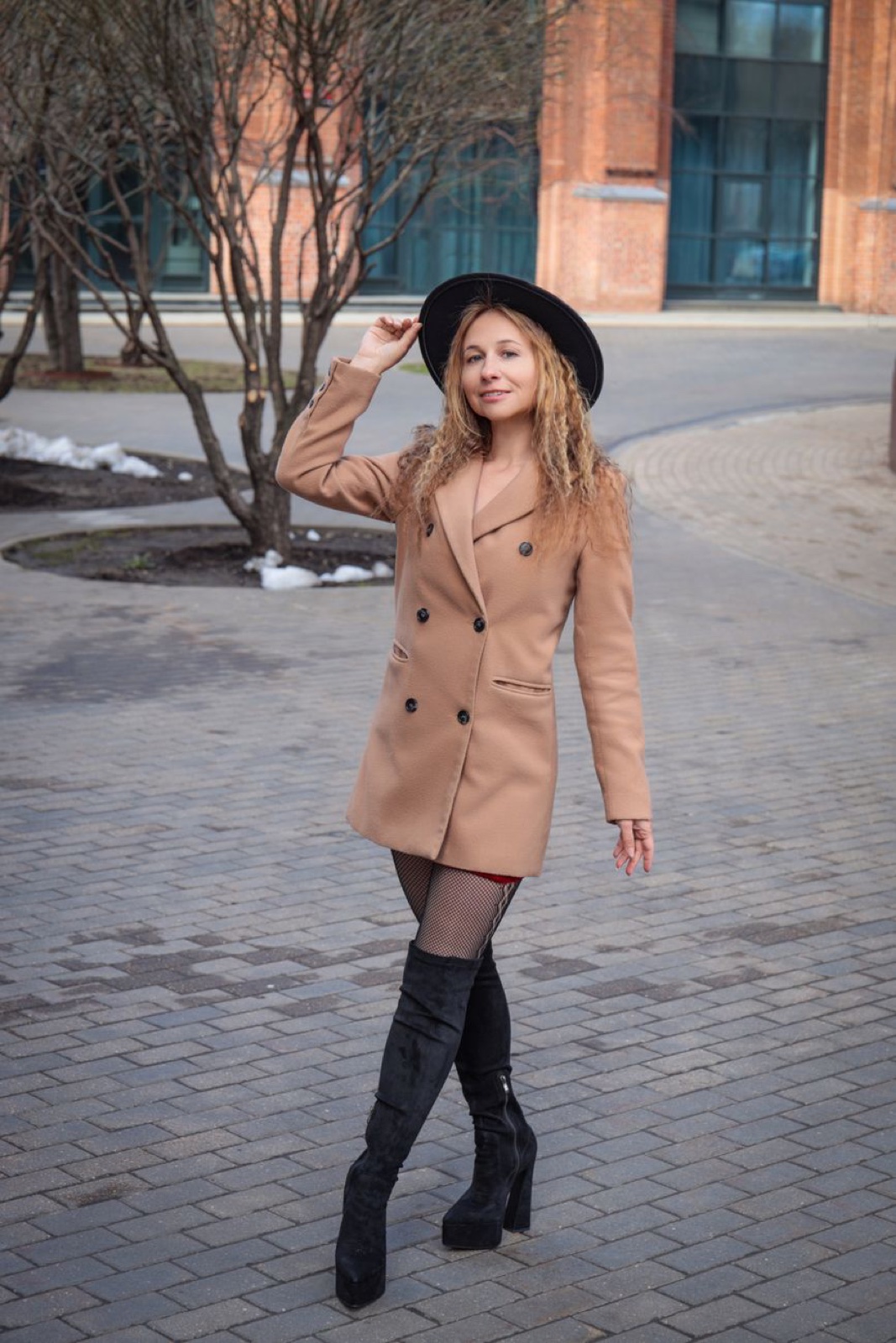A woman in a short double-breasted camel blazer coat, black wide-brim hat and black boots posing on grey paving stones with patches of snow behind her