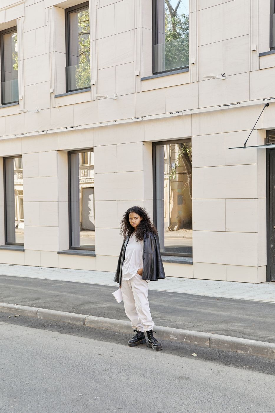 A woman in a white tee, off-white pants and black boots with a black leather jacket over her shoulders, standing on a curb by a pale stone building