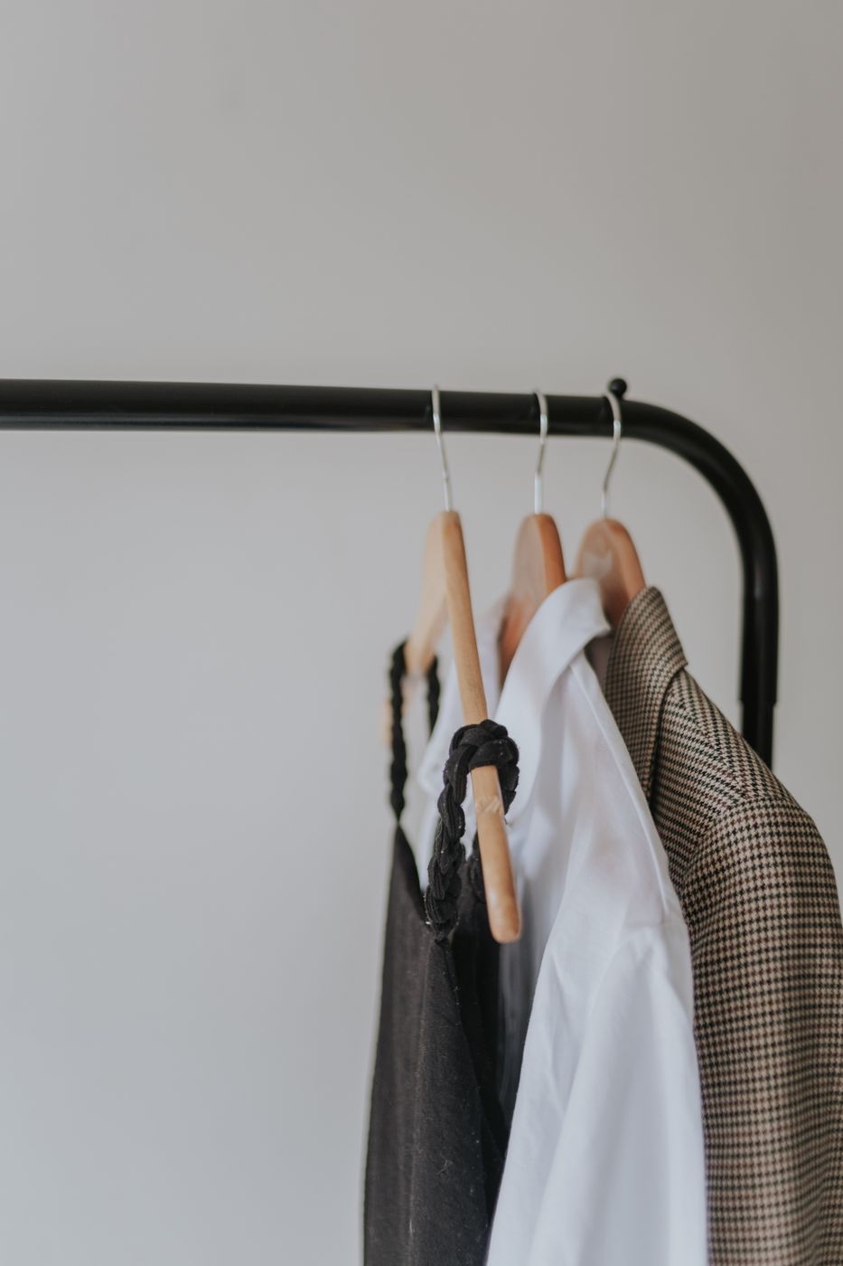 A white shirt on a wooden hanger between a black slip top and a houndstooth blazer on a black clothing rack