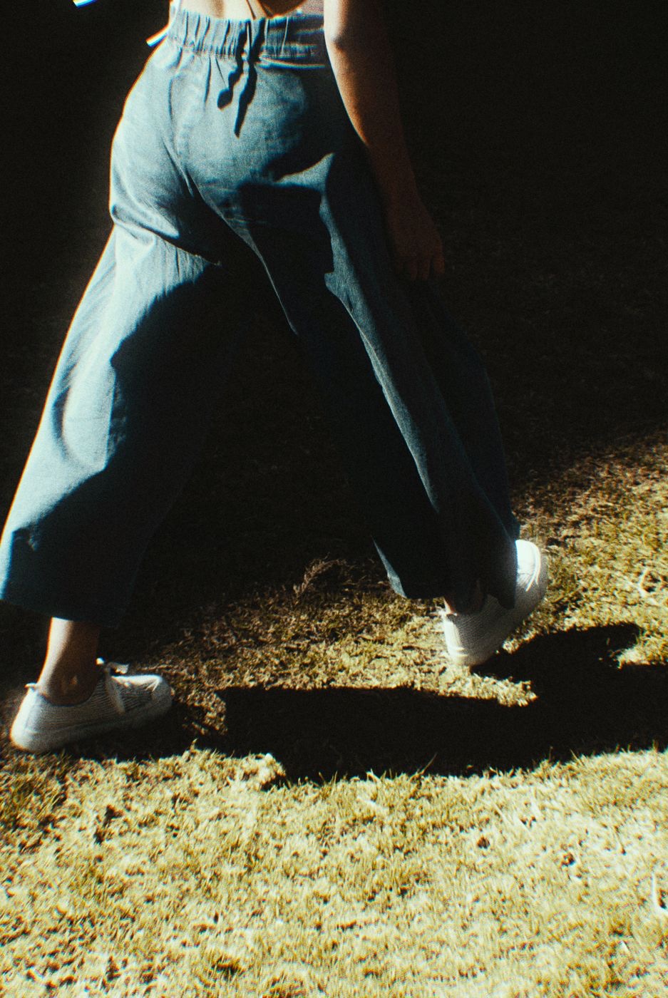 A person photographed from behind at night walking across grass in cropped wide-leg jeans, the outseam curving away from the ankle
