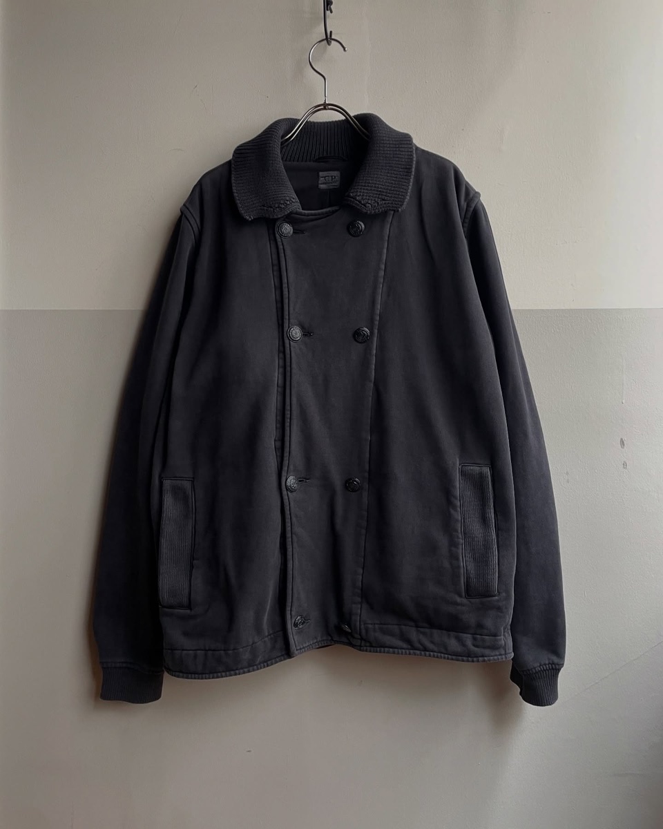 A faded charcoal C.P. Company double-breasted pea coat with a ribbed knit collar hanging on a wire hanger against a plain wall