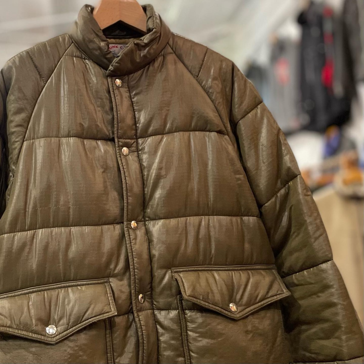 An olive ripstop puffer jacket with pearl snaps and a worn sheen hanging on a wooden hanger in a secondhand clothing shop