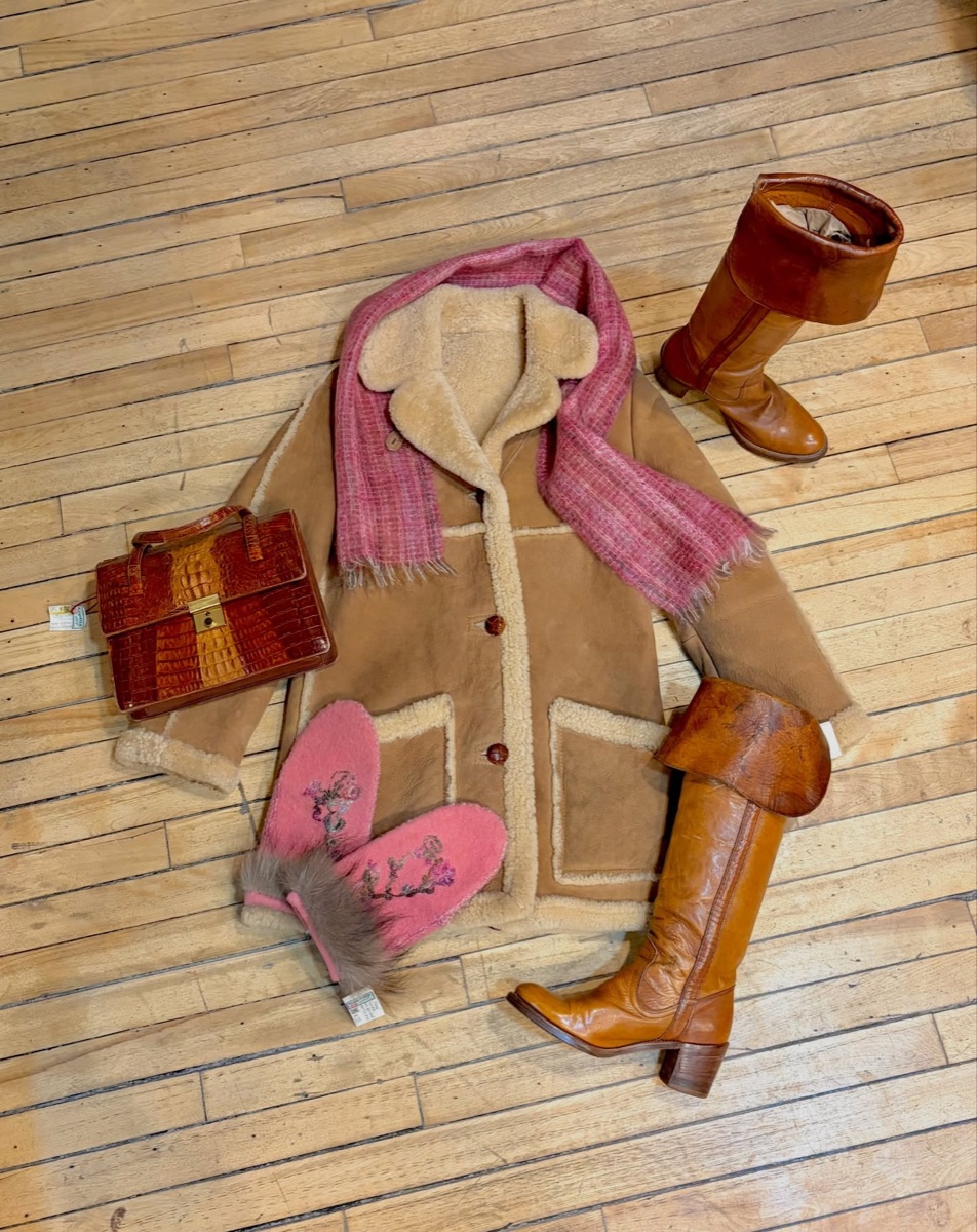 A tan shearling coat with cream fleece collar laid flat on a wooden floor beside cognac leather riding boots, a crocodile-stamped bag and a pink mohair scarf, price tags attached