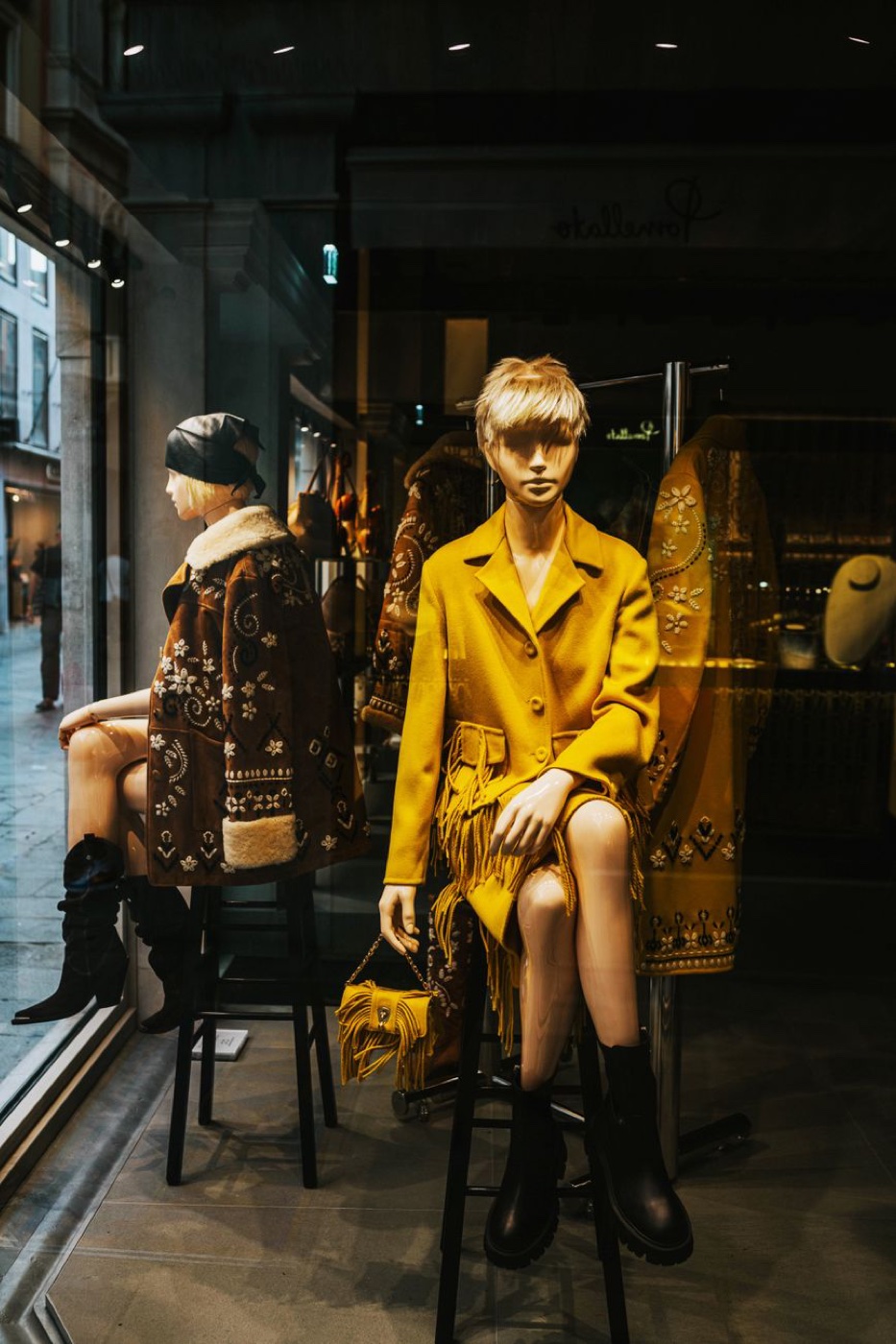 Two seated mannequins in a shop window wearing embroidered brown shearling and mustard wool coats, with boots and a fringed bag
