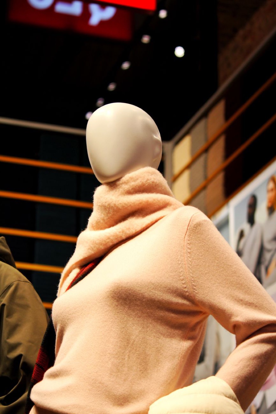 A faceless store mannequin in a peach turtleneck jumper and matching scarf, photographed from below against warm shop lighting