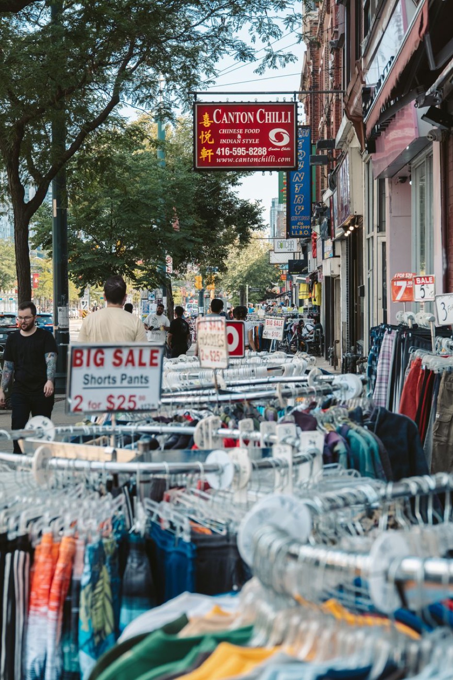 Sidewalk clothing racks outside a shop with a handwritten-style sign reading Big Sale Shorts Pants 2 for 25 dollars