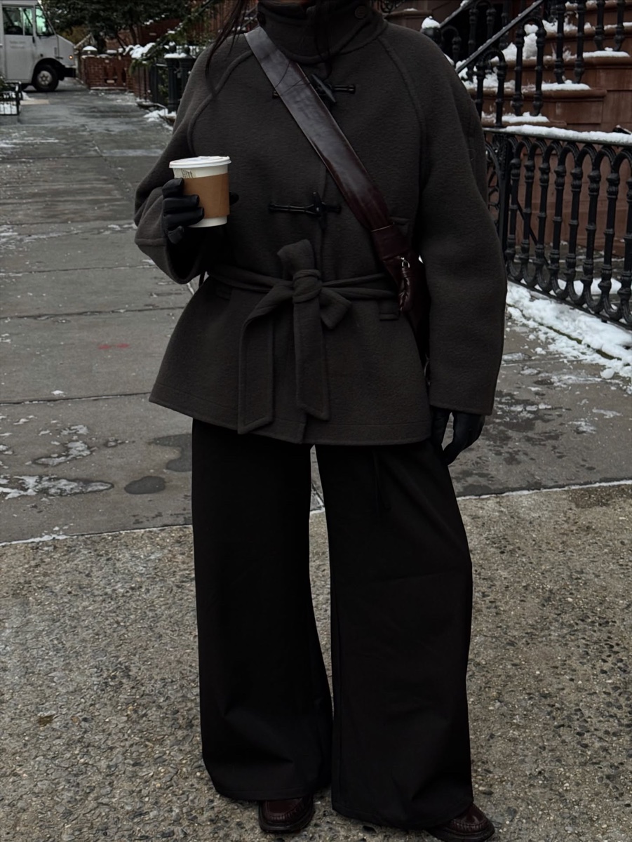 Person on a snowy New York sidewalk in a dark brown belted wool toggle coat, black leather gloves, holding a paper coffee cup, brownstone railings behind