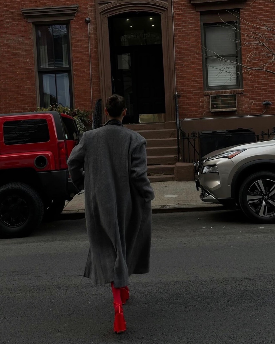 Person seen from behind crossing a Brooklyn street in a long grey wool coat with padded shoulders and red boots, brick townhouses and parked cars ahead