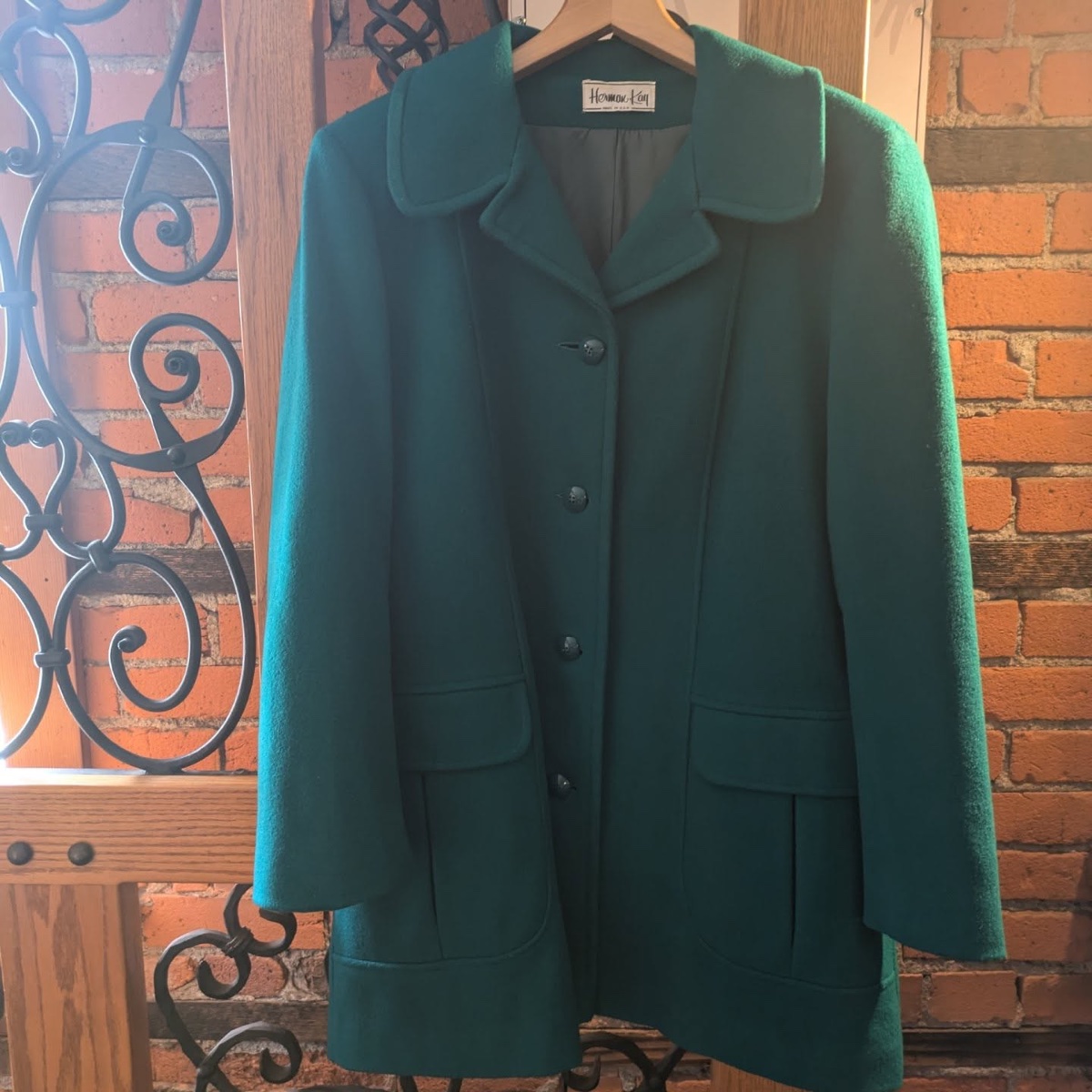 Emerald green vintage wool coat on a wooden hanger against a brick wall, Herman Kay label visible at the neck, flap pockets and covered buttons