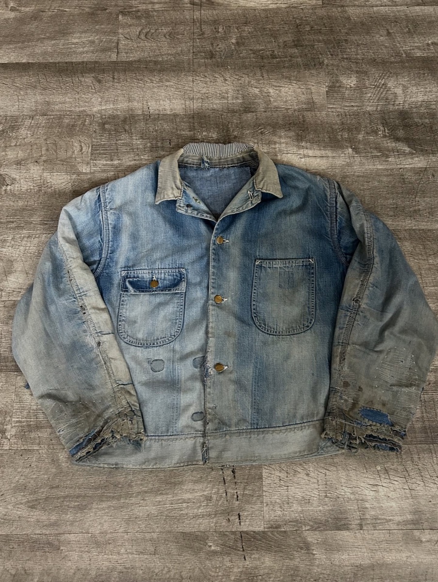 A faded blue denim chore jacket laid flat on gray wood, cuffs frayed to threads and a striped corduroy collar worn pale