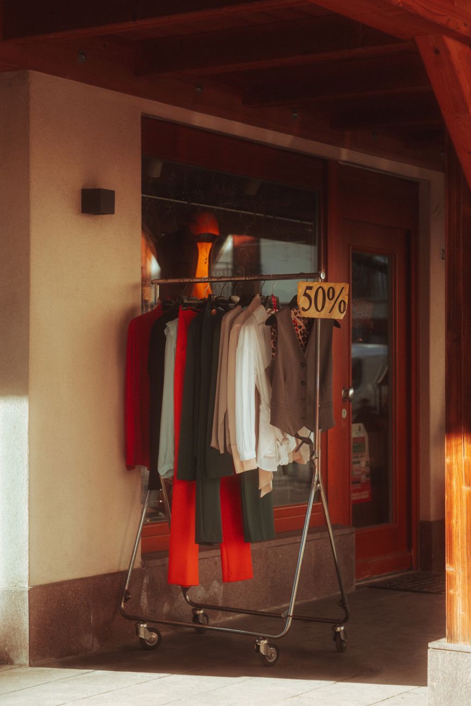 A wheeled clothing rail parked outside a shop doorway in late sun, hung with red, olive and cream pieces, a handwritten cardboard sign reading 50% clipped to the end