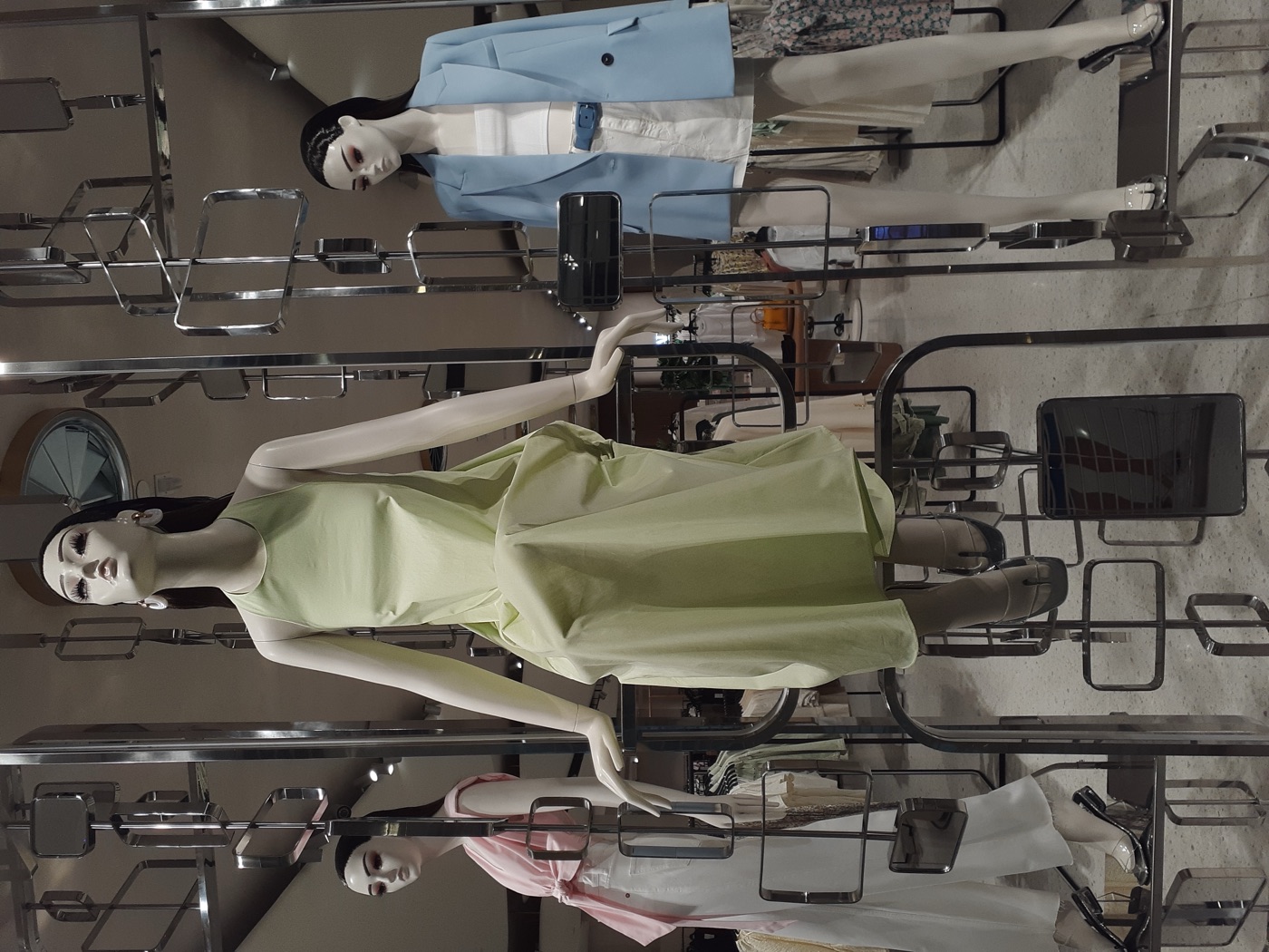 Mannequins in a mall window styled in a pale green dress, a pink top with white trousers and a light blue blazer with white shorts, chrome display frames around them