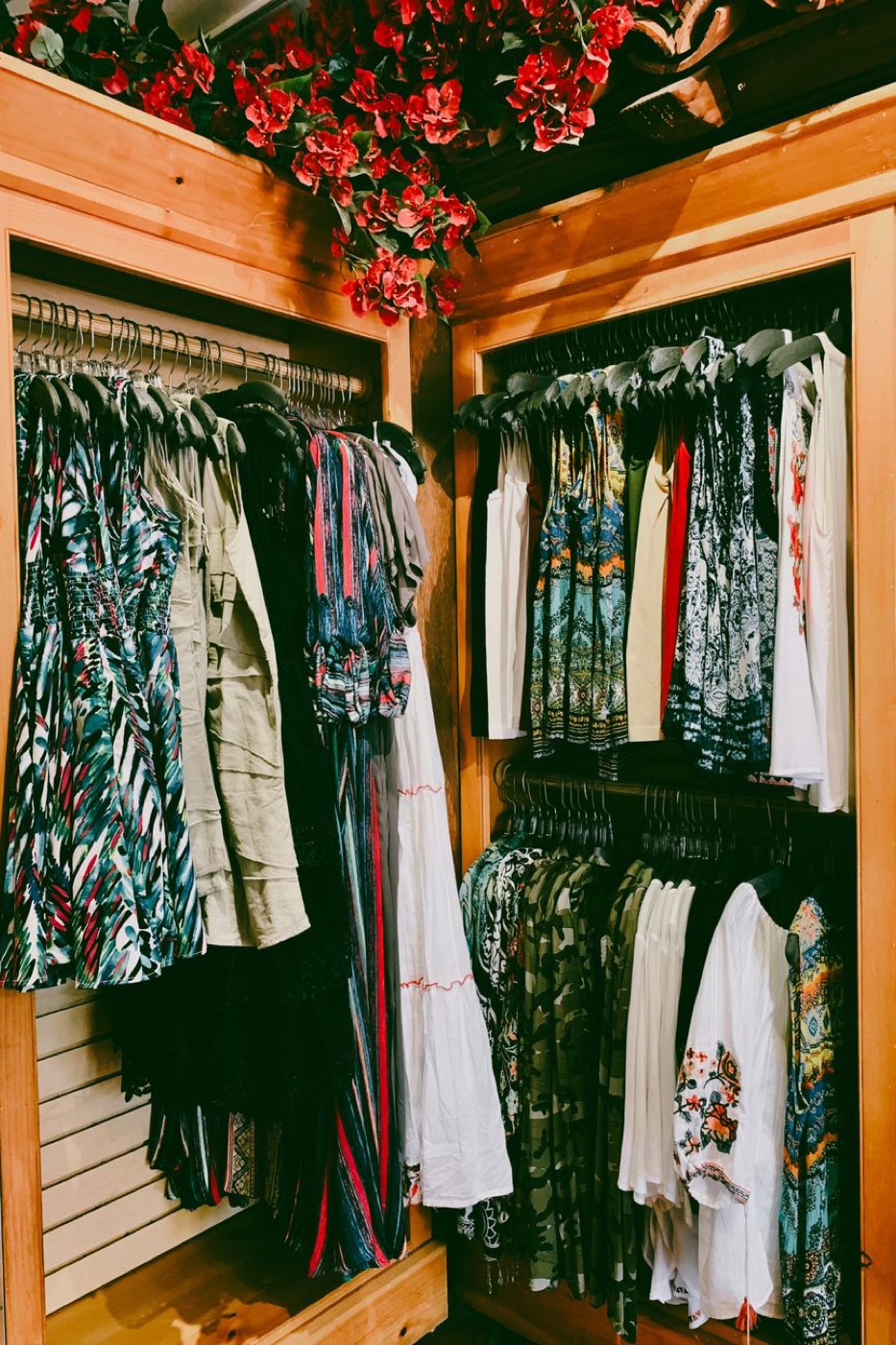 Two wooden alcoves of a boutique packed with printed summer dresses and light cotton tops on black and silver hangers, artificial red flowers along the top rail