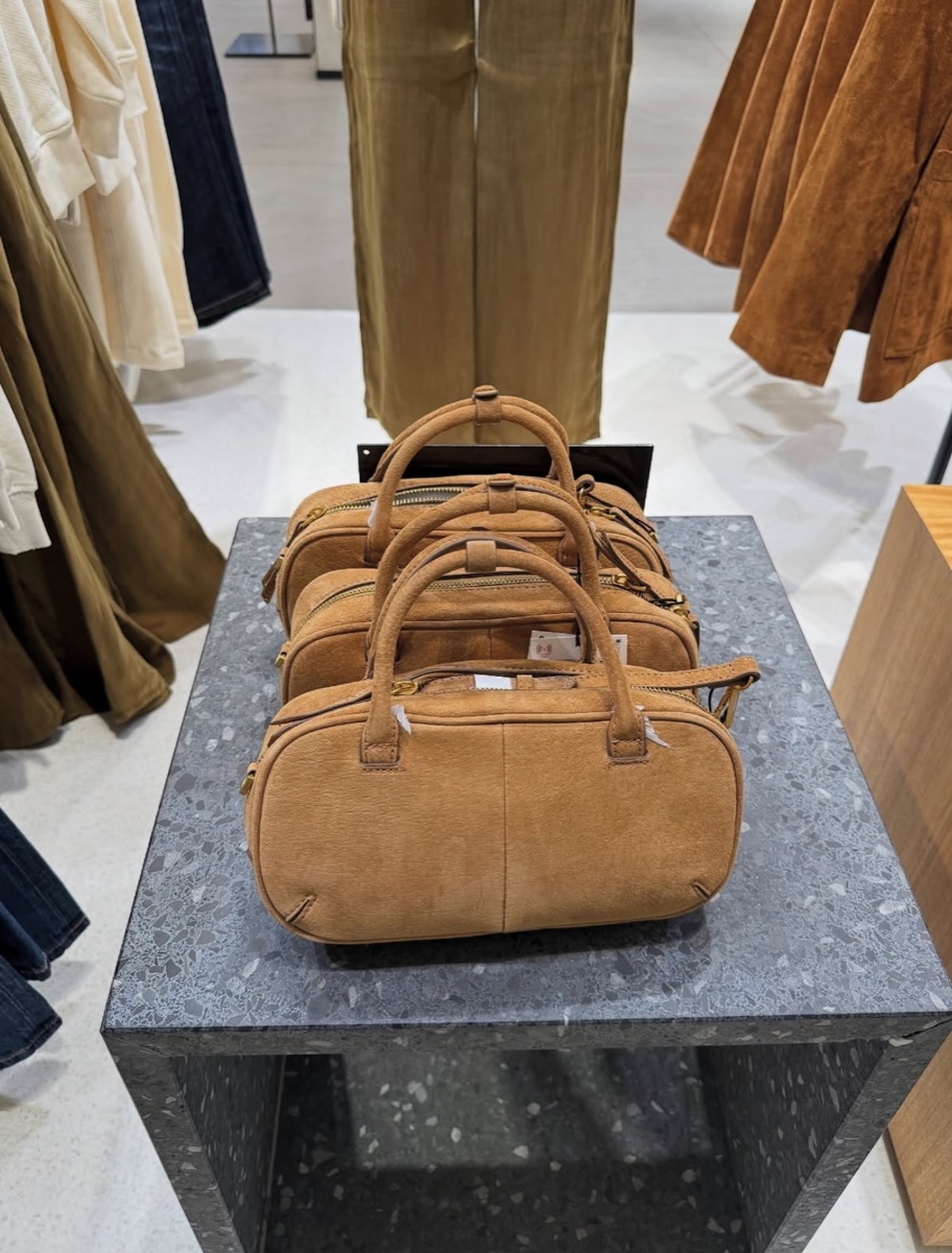 Three tan split-suede mini bowling bags with paper tags lined up on a grey terrazzo display block in a Zara store, suede skirts on the rail behind