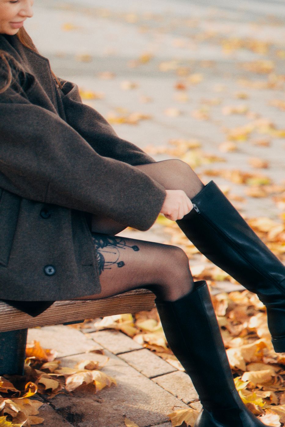A woman on a park bench zipping tall black boots over patterned sheer tights among fallen leaves